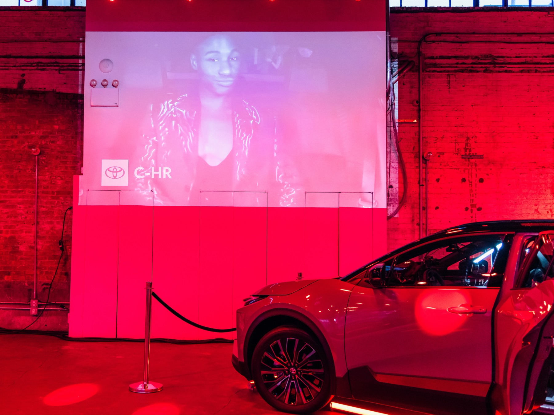 The Toyota C-HR staged on the activation floor, door open, with the in-car camera feed projected onto the venue wall behind it under red lighting and a disco ball overhead